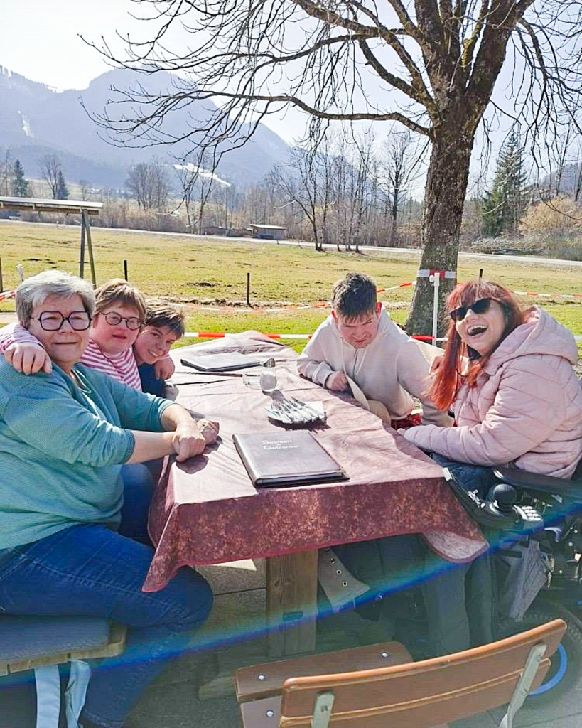 Vier Mitglieder der WG mit Herz und einer Betreuungsperson an einem Picknicktisch im Außenbereich bei einem Ausflug